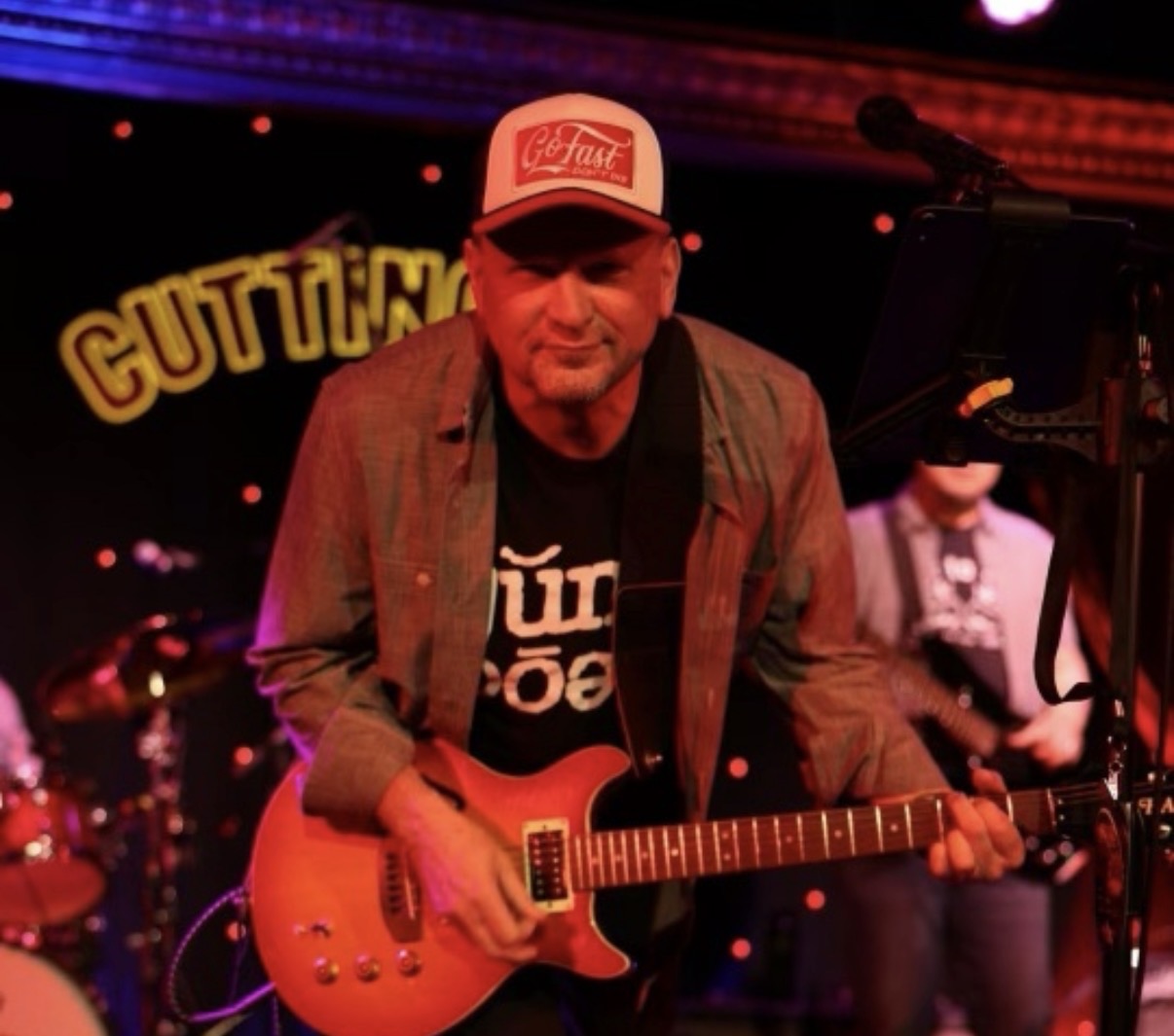 John playing electric guitar under red stage lighting, with a drummer behind him